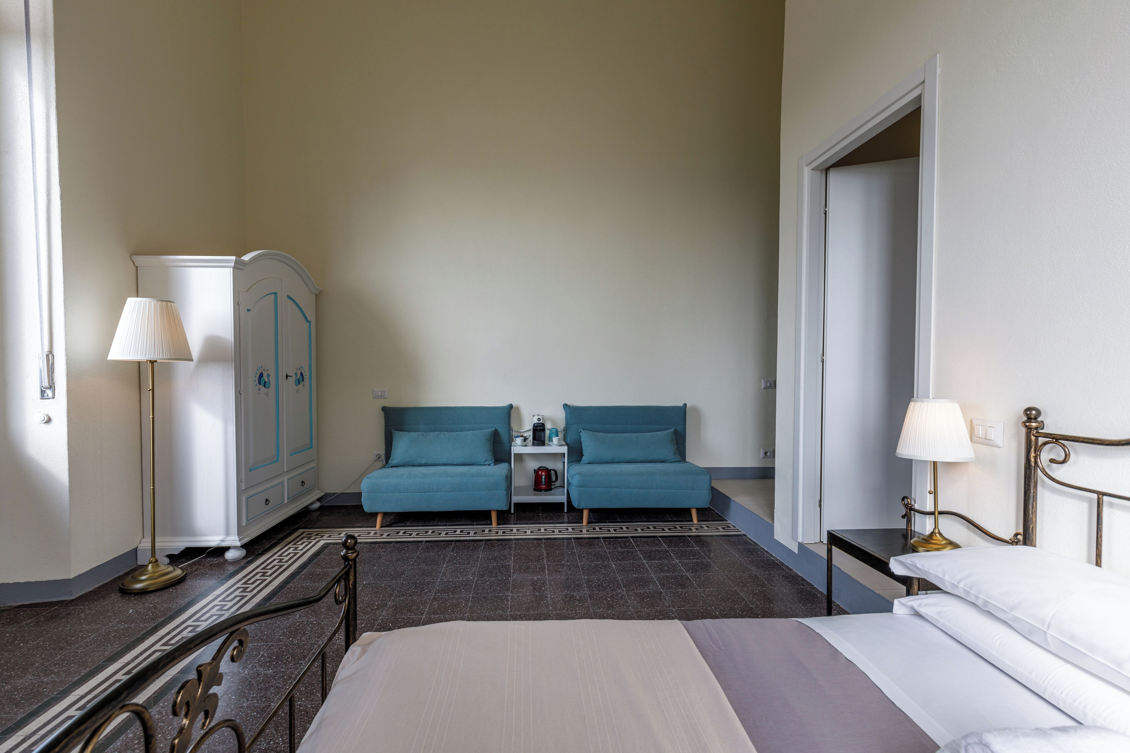 A hotel room with a bed, two blue sofas, a white wardrobe, a bedside table with a lamp, and a floor lamp.