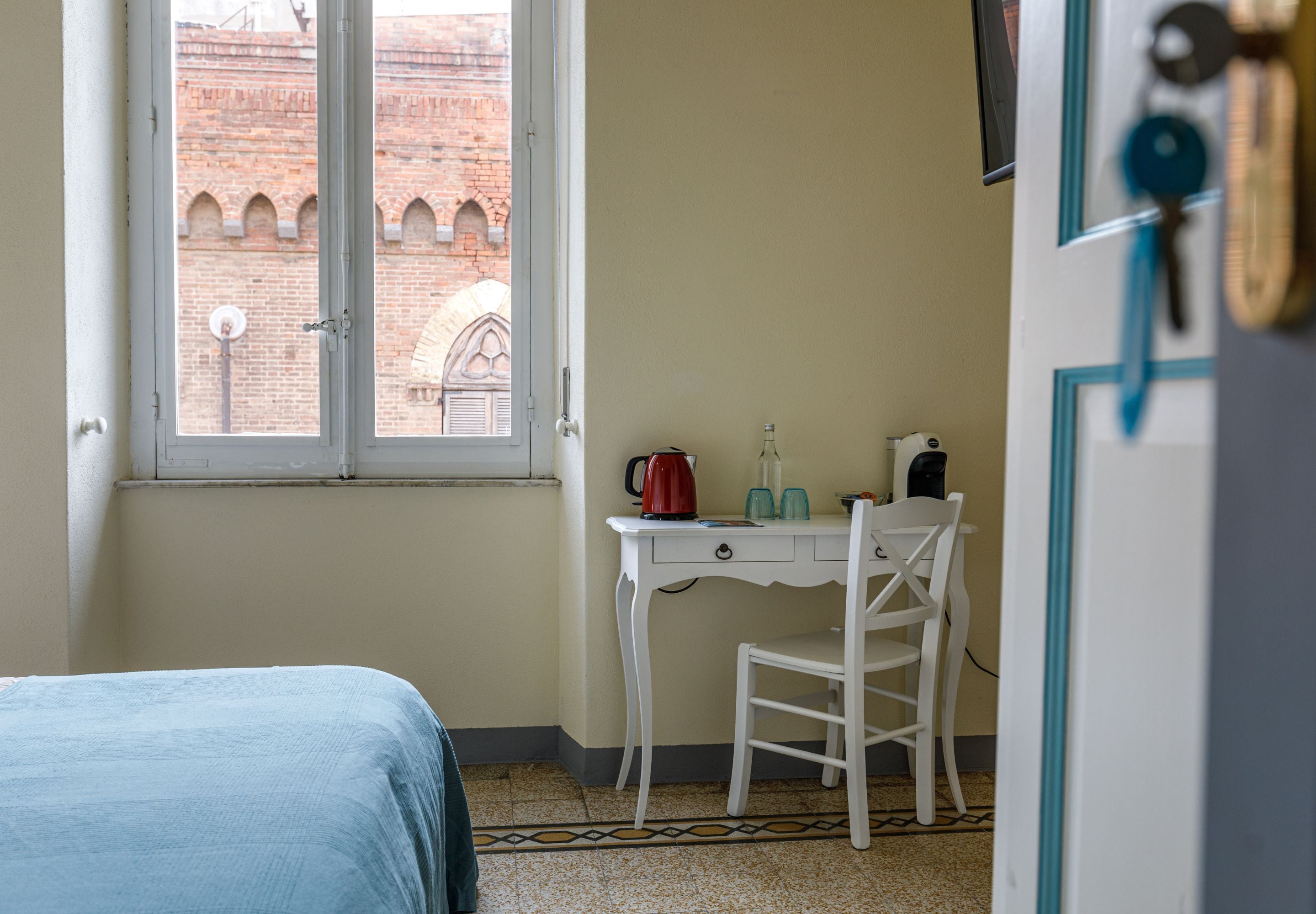 Ein Zimmer mit einem Bett, einem Schreibtisch mit Stuhl, einem Fenster mit Blick auf ein Ziegelgebäude und einer Tür, an der ein Schlüssel hängt.