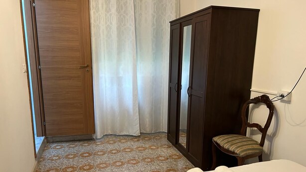 A hotel room with a bed, a wooden wardrobe, a chair, and a curtain. - VENICE NIGHT 25 DOUBLE ROOM  A (Mestre)