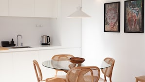 Cozinha moderna com armários brancos, pia de aço inoxidável e cafeteira. Há uma mesa redonda de vidro com quatro cadeiras de vime. Duas obras de arte emolduradas estão penduradas na parede.