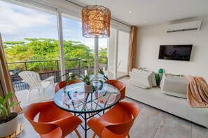 Relax in this stylish living room with a glass-top dining table, comfortable sofa, and large windows offering a view of lush greenery.