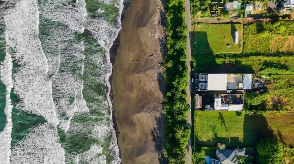 Vue aérienne d’une propriété située sur la côte, avec une plage de sable, une zone herbeuse et quelques bâtiments.