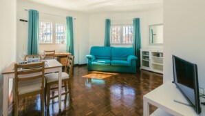 A living room with a wooden dining table, chairs, a teal sofa, a TV, and a window with curtains. - Madrid Rio Perfect location for exploring Madrid! Modern 1-bed flat sleeps 2. (Madrid)