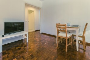 Ein Wohnzimmer mit Flachbildfernseher auf weißem Standfuß, einem Holzessttisch mit zwei Stühlen und Holzboden.