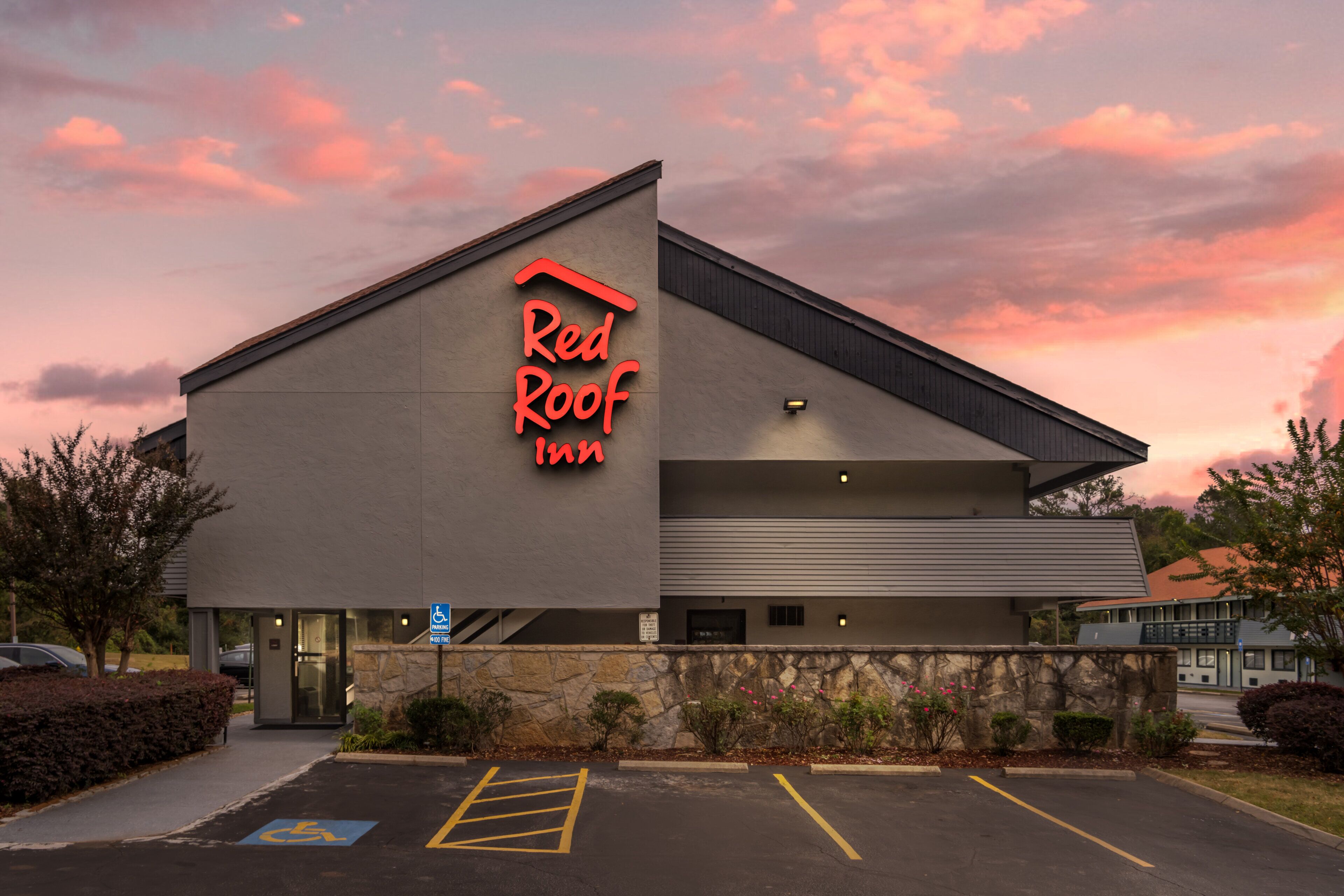 Un hôtel Red Roof Inn doté d’une architecture moderne, avec une enseigne au néon bien visible, un parking accessible aux personnes handicapées et une façade en pierre.