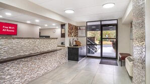 A reception area with a tiled wall, a reception desk, a digital screen displaying "Free Wi-Fi All Rooms," and a view of the outdoors through a glass door.