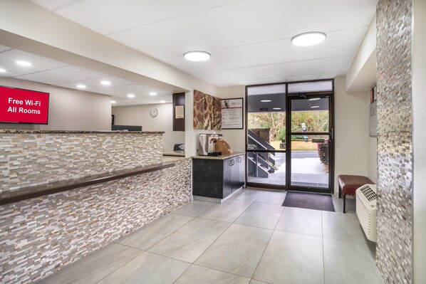 A reception area with a tiled wall, a reception desk, a digital screen displaying "Free Wi-Fi All Rooms," and a view of the outdoors through a glass door.