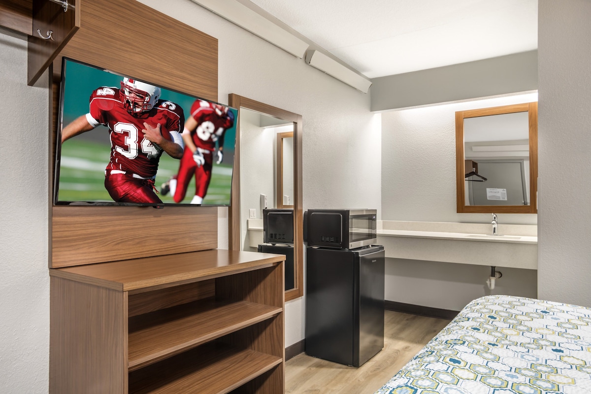A hotel room with a flat-screen TV mounted on the wall, a wooden TV stand, a black refrigerator, a microwave, a wooden bed, and a mirror with a wooden frame.