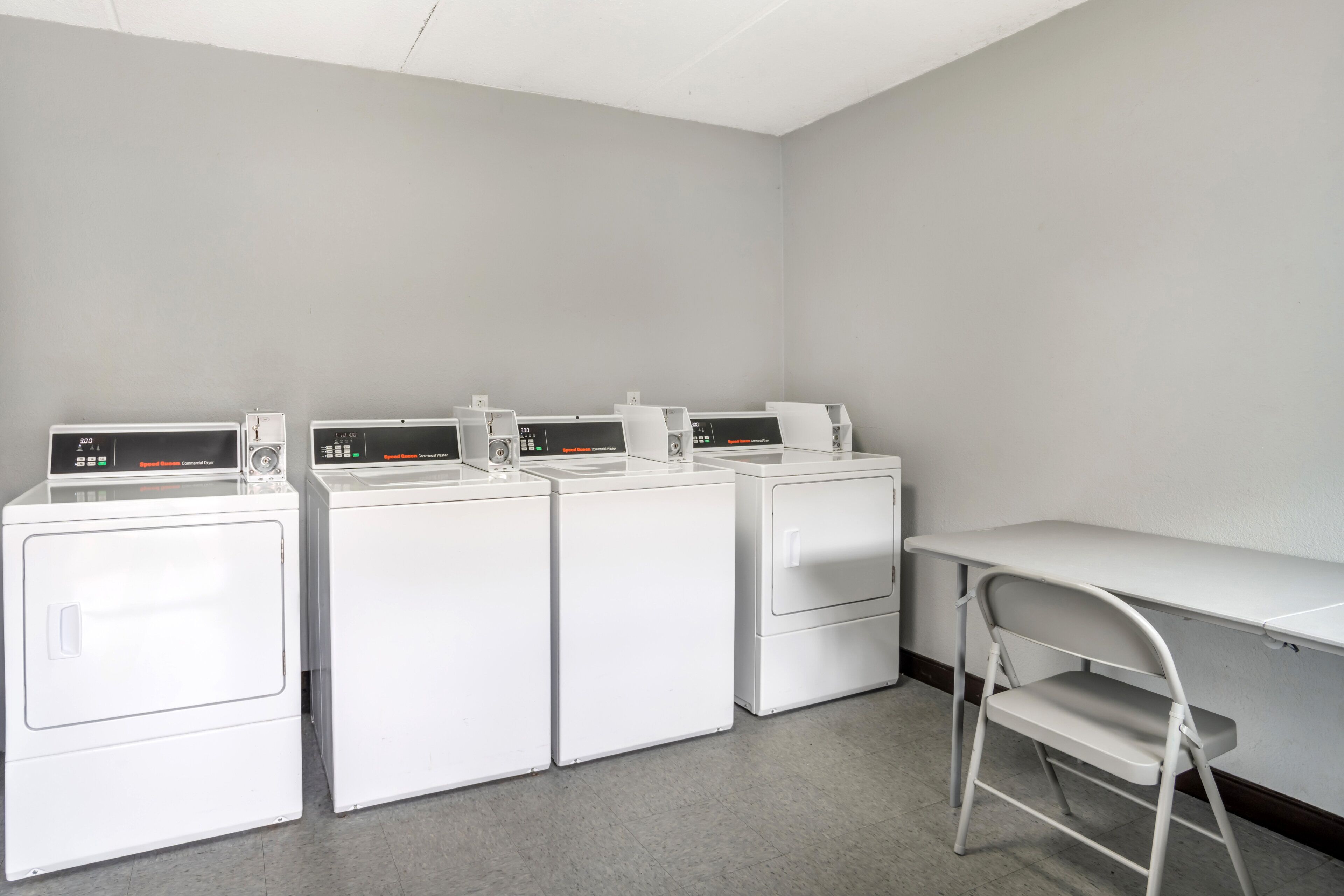 A laundry room with four white commercial washers and dryers, a folding table, and a chair.