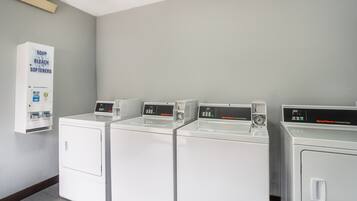 A laundry facility with four white washing machines and a soap, bleach, and softeners dispenser.