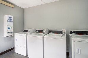 A laundry facility with four white washing machines and a soap, bleach, and softeners dispenser. - HomeTowne Studios by Red Roof Greenville (Greenville)