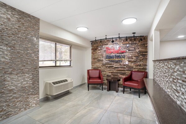 Un espace d’accueil avec un mur en pierre, deux fauteuils en cuir rouge, une petite table et une fenêtre avec des stores.