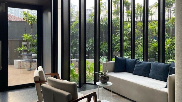 A modern living room with a sofa, chairs, and a small table. Large windows offer a view of greenery outside. - Onze22 NR (São Paulo)