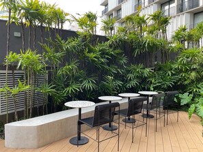 An outdoor patio area with round tables and chairs, surrounded by lush greenery and a modern building in the background. - Onze22 NR (São Paulo)