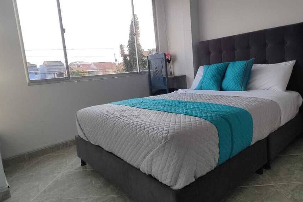 Un dormitorio con cama, mesita de noche, ventana y vistas a edificios.