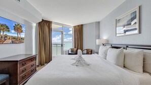 Een hotelkamer met een groot bed, een bureau met lades, een stoel, een balkon met gordijnen en een schilderij aan de muur.