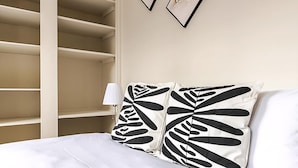 Un lit avec une literie blanche et deux coussins à motifs noir et blanc, une lampe de chevet et une étagère blanche adossée à un mur beige.