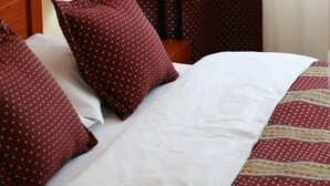 A neatly made bed with white and patterned bedding, a wooden headboard, a lamp on a bedside table, and red curtains with a polka dot pattern. - Hotel Apollonia (Pristina)