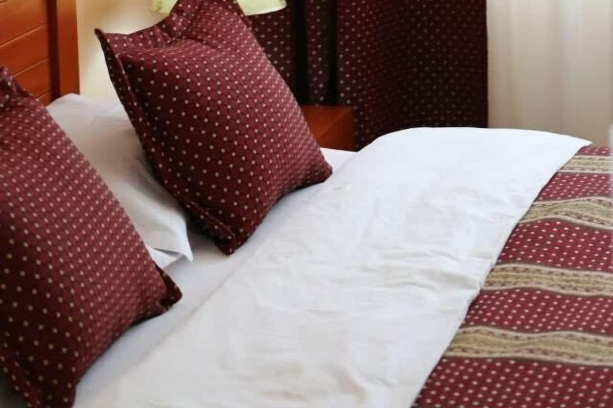 A neatly made bed with white and patterned bedding, a wooden headboard, a lamp on a bedside table, and red curtains with a polka dot pattern.