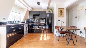 A modern kitchen with a dining area, featuring stainless steel appliances, a wooden dining table, and a large window providing natural light. - Stylish and Sleek Kensington Apartment (London)