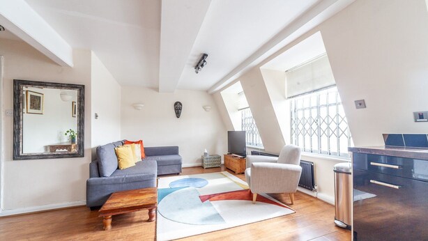 A modern apartment with a loft-style ceiling, a grey sofa, a white armchair, a wooden coffee table, and a kitchen area with a black countertop and cabinets. - Stylish and Sleek Kensington Apartment (London)