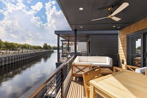 A deck with a view of a canal, a white sofa, wooden chairs, and a table.