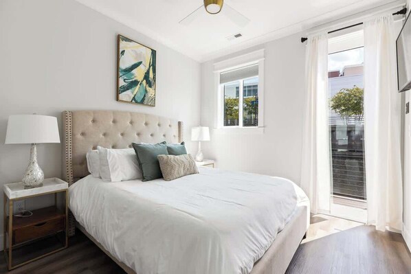 A bedroom with a large bed, two bedside tables, a ceiling fan, a window with curtains, and a painting on the wall.