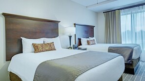 A hotel room with two beds, a nightstand, a lamp, and a window with curtains.