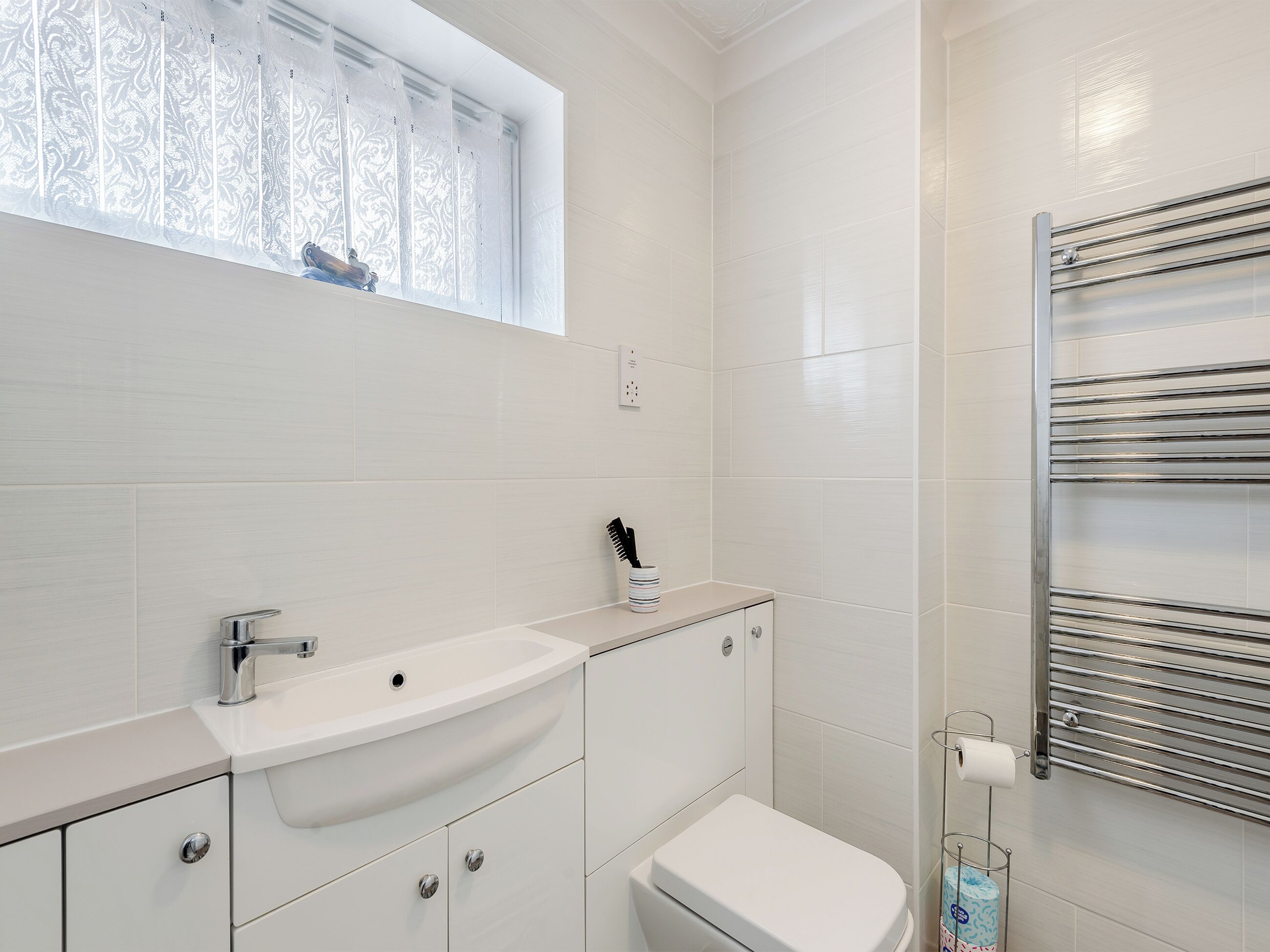 Une salle de bain moderne avec un lavabo, un meuble-lavabo et des toilettes, le tout de couleur blanche. Il y a une fenêtre avec du verre dépoli et un porte-serviettes fixé au mur.