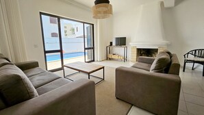 Ein großzügiger Wohnraum mit einer Sofagarnitur, einem Couchtisch und einem Kamin. Ein Fernseher steht auf einem Schrank, und durch die Glastüren ist ein Pool zu sehen.