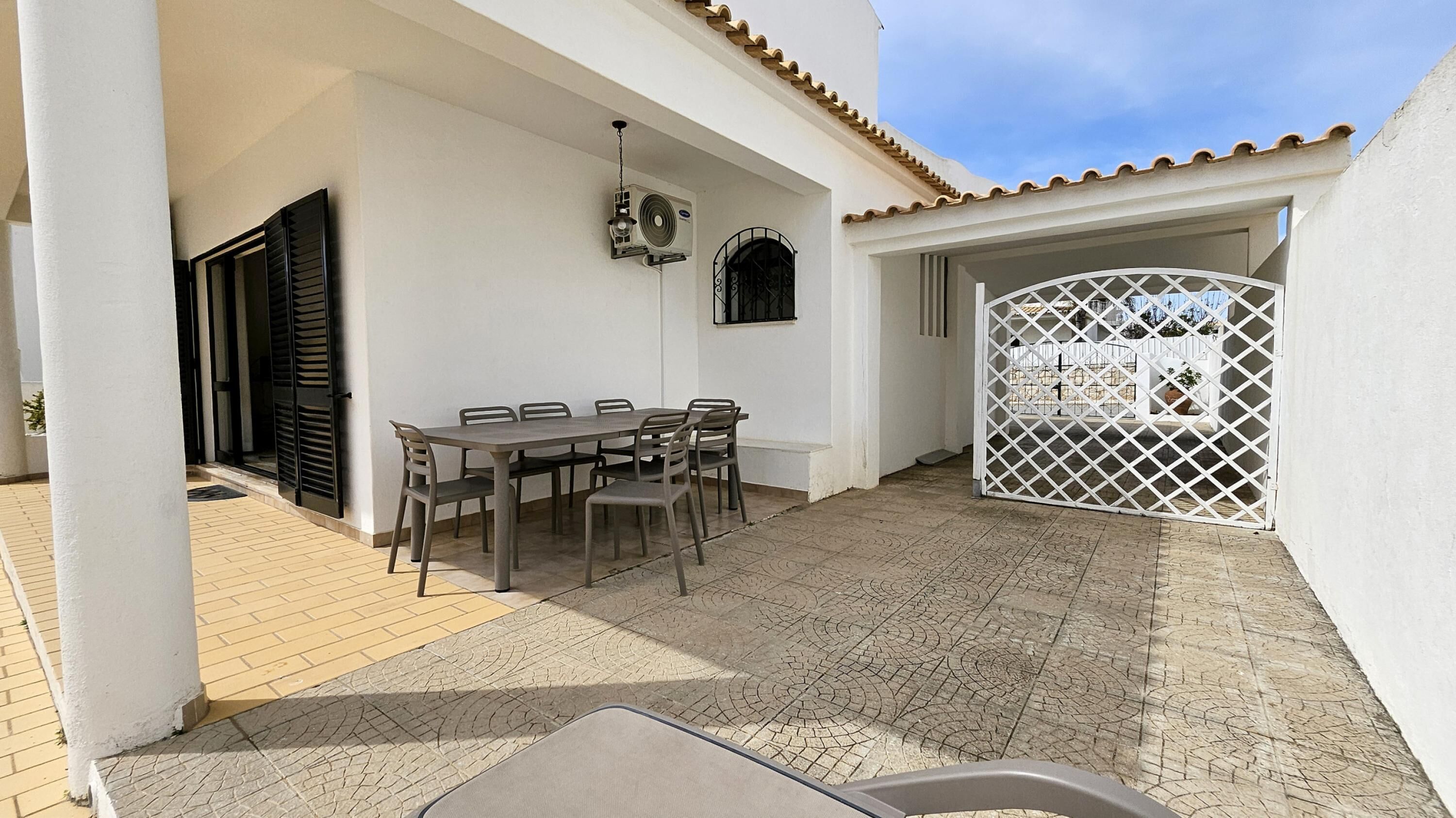 A covered patio with a table and chairs, an air conditioning unit, and a white gate.