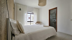 Ein Schlafzimmer mit einem großen Bett, einem Korbstuhl, einem Badezimmer mit Waschbecken und Spiegel sowie einer Tür zu einem anderen Raum.