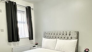 A bedroom with a bed, white curtains, a radiator, a nightstand with a plant, and a grey headboard.