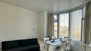 A modern living room with a dark green sofa, a white dining table with chairs, and a large window with curtains.
