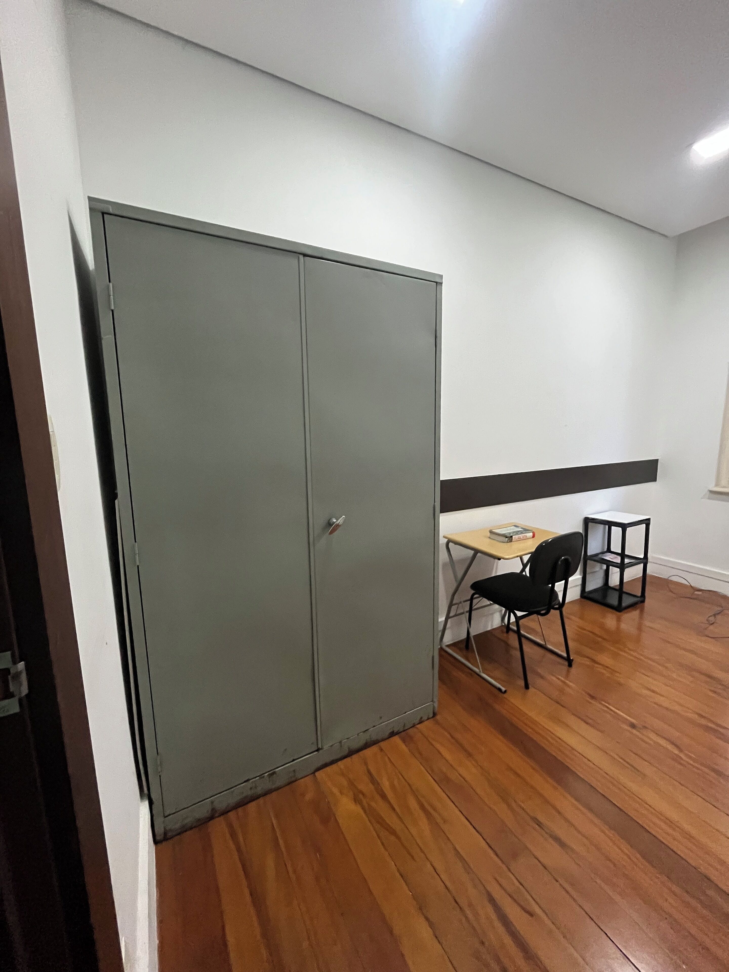 A room with a grey wardrobe, a small wooden table with a chair, and a black shelf unit.