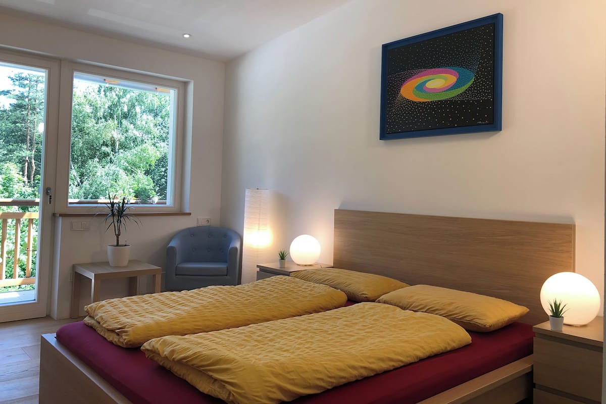 A bedroom with a double bed, a chair, a bedside table, a lamp, and a window with a view of trees.
