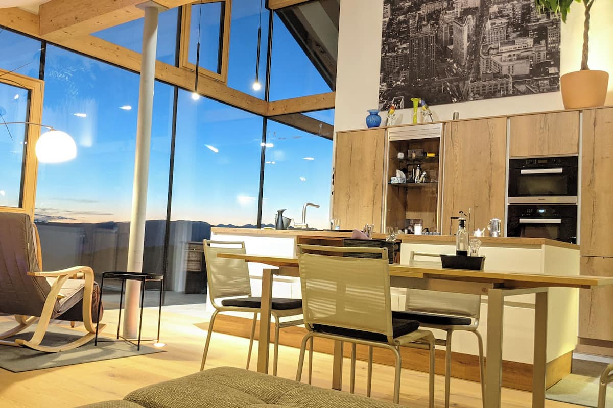 A modern kitchen with large windows, wooden cabinets, and a dining area with chairs and a table.