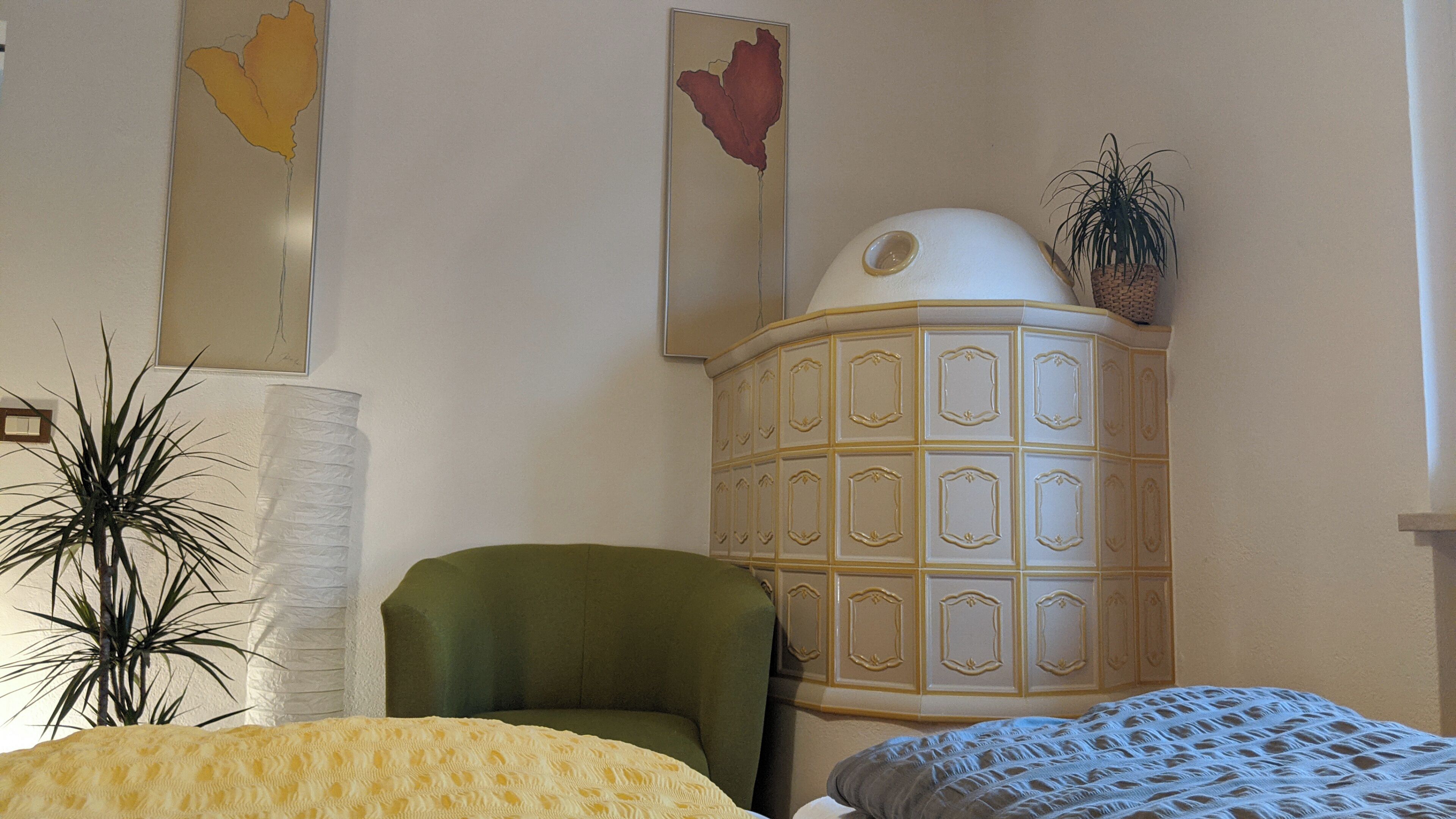 Una stanza con una poltrona verde, un armadietto bianco con pannelli decorativi, una coperta giallo fantasia e una pianta in vaso.