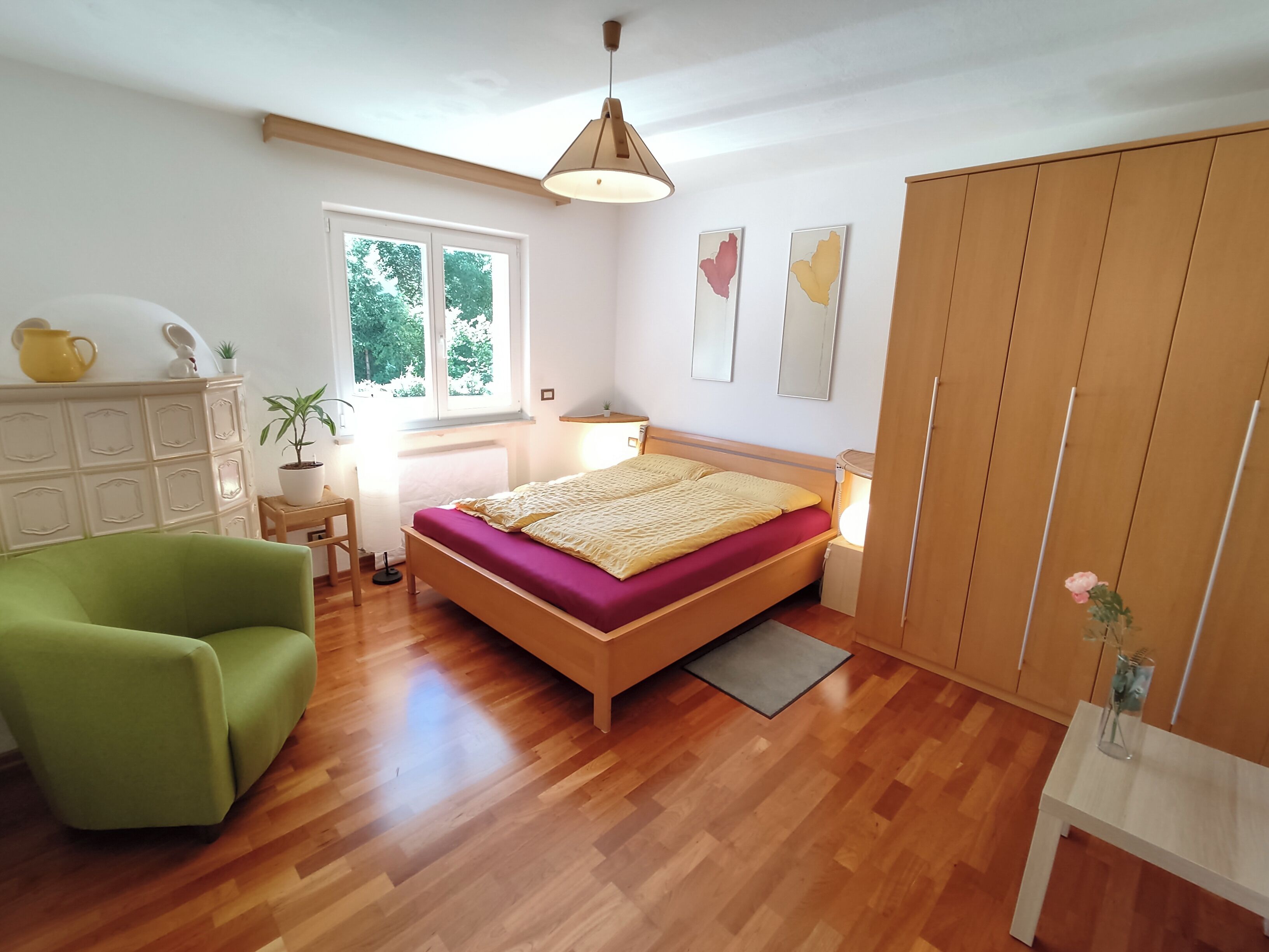 A bedroom with a wooden bed, a green armchair, a dresser, a window, and a wardrobe.