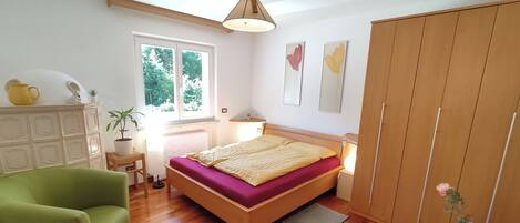 A bedroom with a wooden bed, a green armchair, a dresser, a window, and a wardrobe.