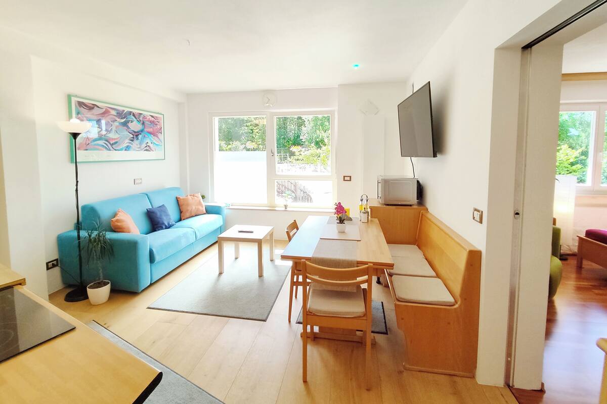 A modern living room with a blue sofa, a dining table with chairs, a flat-screen TV mounted on the wall, and large windows providing natural light.