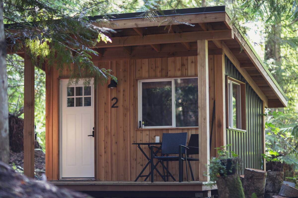 Una pequeña cabaña de madera con una puerta, una ventana y una mesa con dos sillas.