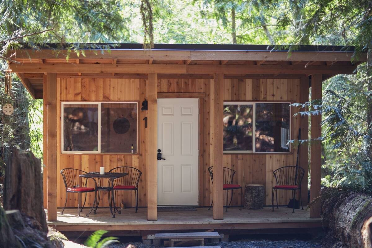 Una cabaña de madera con dos ventanas, una puerta y un pequeño porche con dos sillas y una mesa.