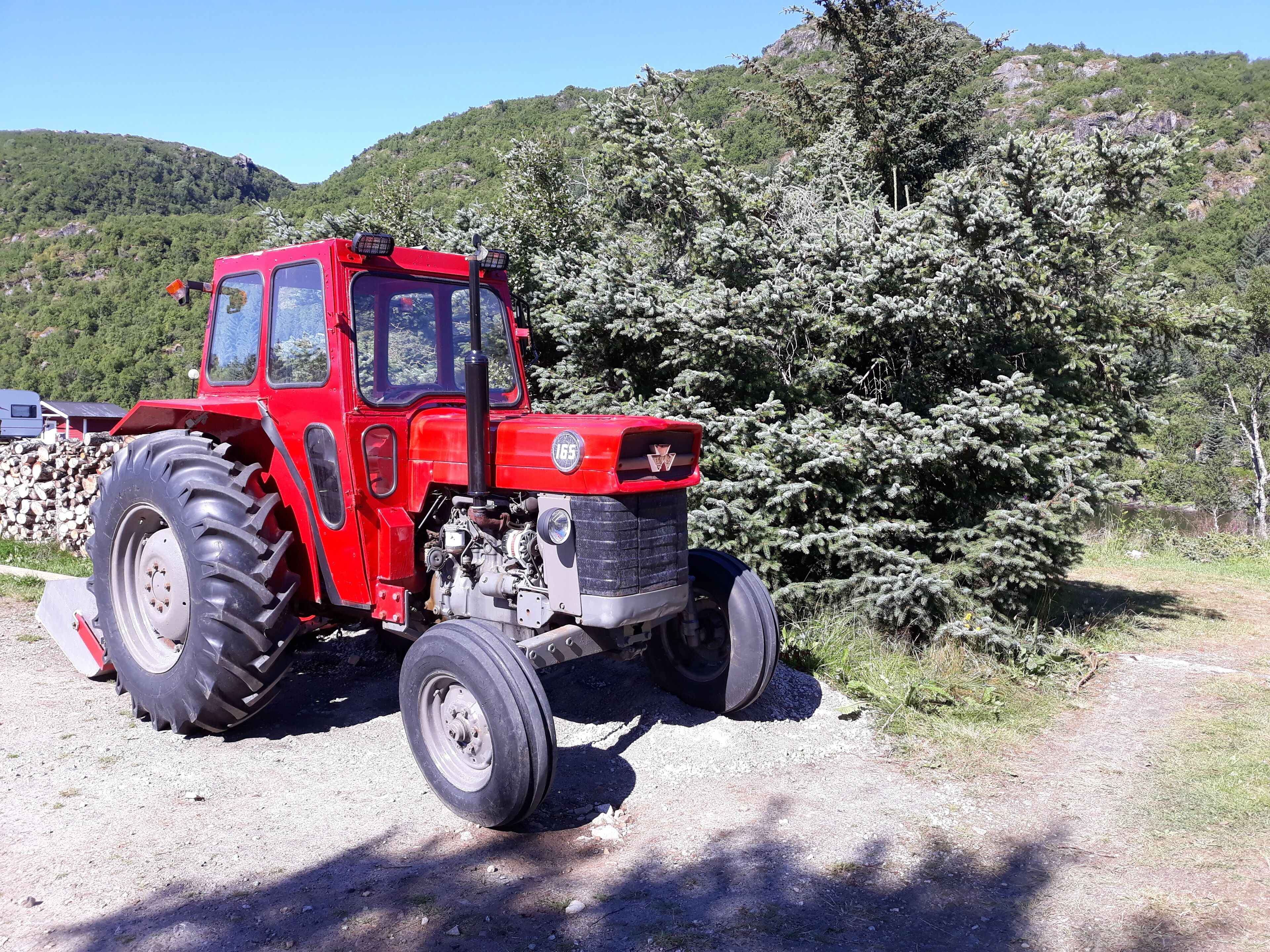 Ein roter Traktor steht auf einem Schotterweg, im Hintergrund ist ein bewaldeter Hügel zu sehen.