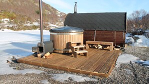 A wooden deck with a hot tub and seating area, surrounded by snow and a mountainous backdrop. - Lofoten Camp Storfjord (Vestvågøy)