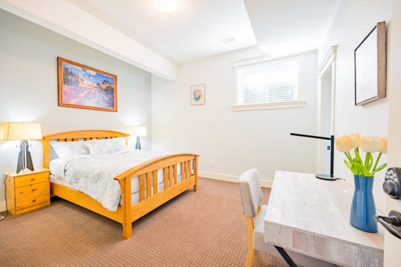 A bedroom with a wooden bed, a nightstand, a desk, a chair, and a window with blinds.