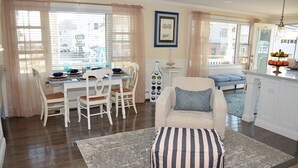 Un coin repas avec une table dressée pour quatre personnes, des chaises blanches, un pouf à rayures et un tapis aux motifs bleus et blancs.
