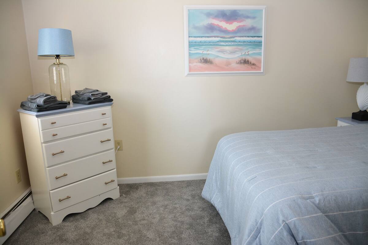 Een slaapkamer met een bed, een dressoir met opgevouwen handdoeken, een lamp en een schilderij van een strand.