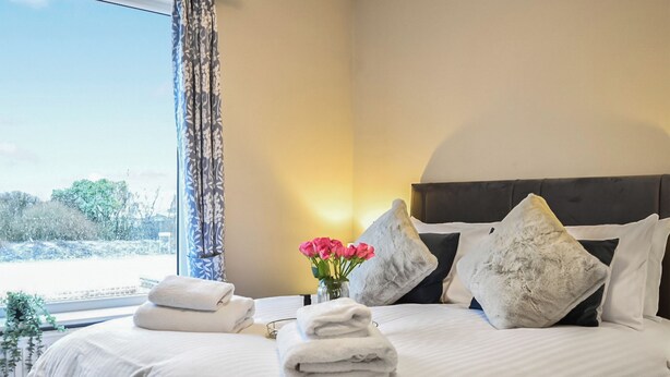 A neatly made bed with white linens and pillows, a vase with pink flowers, and a window with curtains.