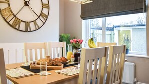 A dining area with a wooden table set for breakfast, a large wall clock, and a window with a view of the outside.